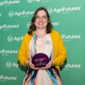 Cheryl McCarthy, AgriFutures Australia researcher of the year. Cheryl is wearing a yellow cardigan and multicolored dress. She is standing in front of a green agrifutures australia sign holding a trophy.