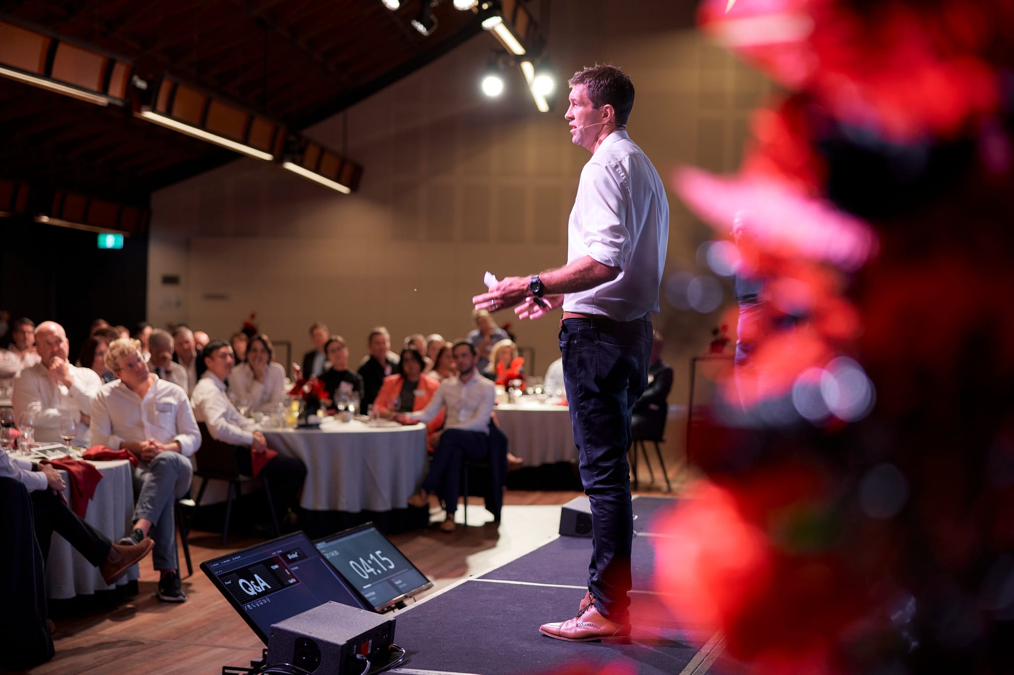 A tall man in a while shirt standing on a stage pitching to a room of people over dinner. 