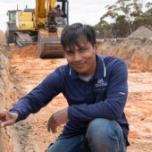 Dr Gaus Azam sitting in a soil pit and pointing at his ruler. He is wearing a DPIRD tshirt and an excavator is in the background